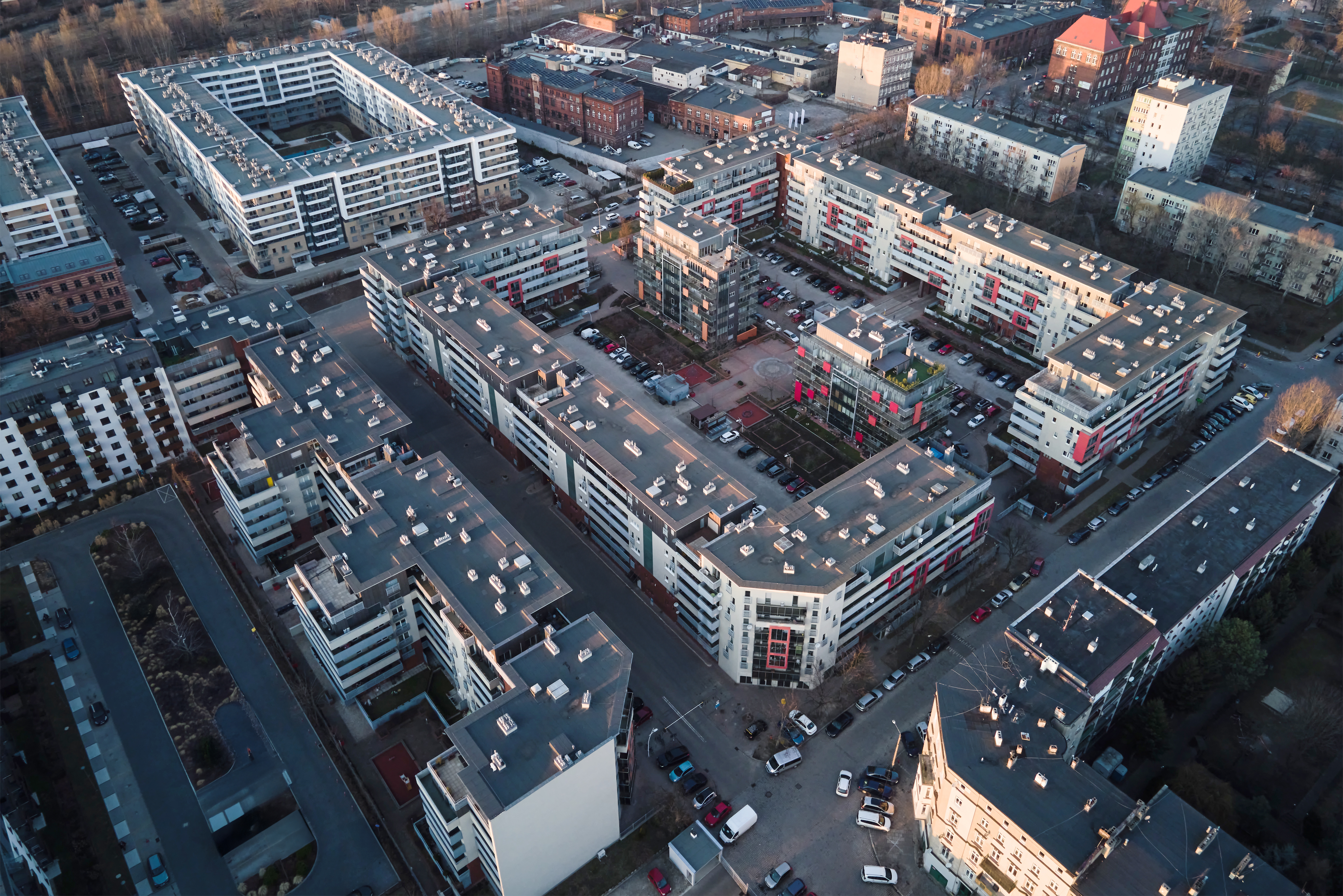 Zdjęcie lotnicze nowoczesnego osiedla mieszkaniowego z wielopiętrowymi blokami o jasnych elewacjach, szarych i czerwonych akcentach. Między budynkami są dziedzińce, parkingi.