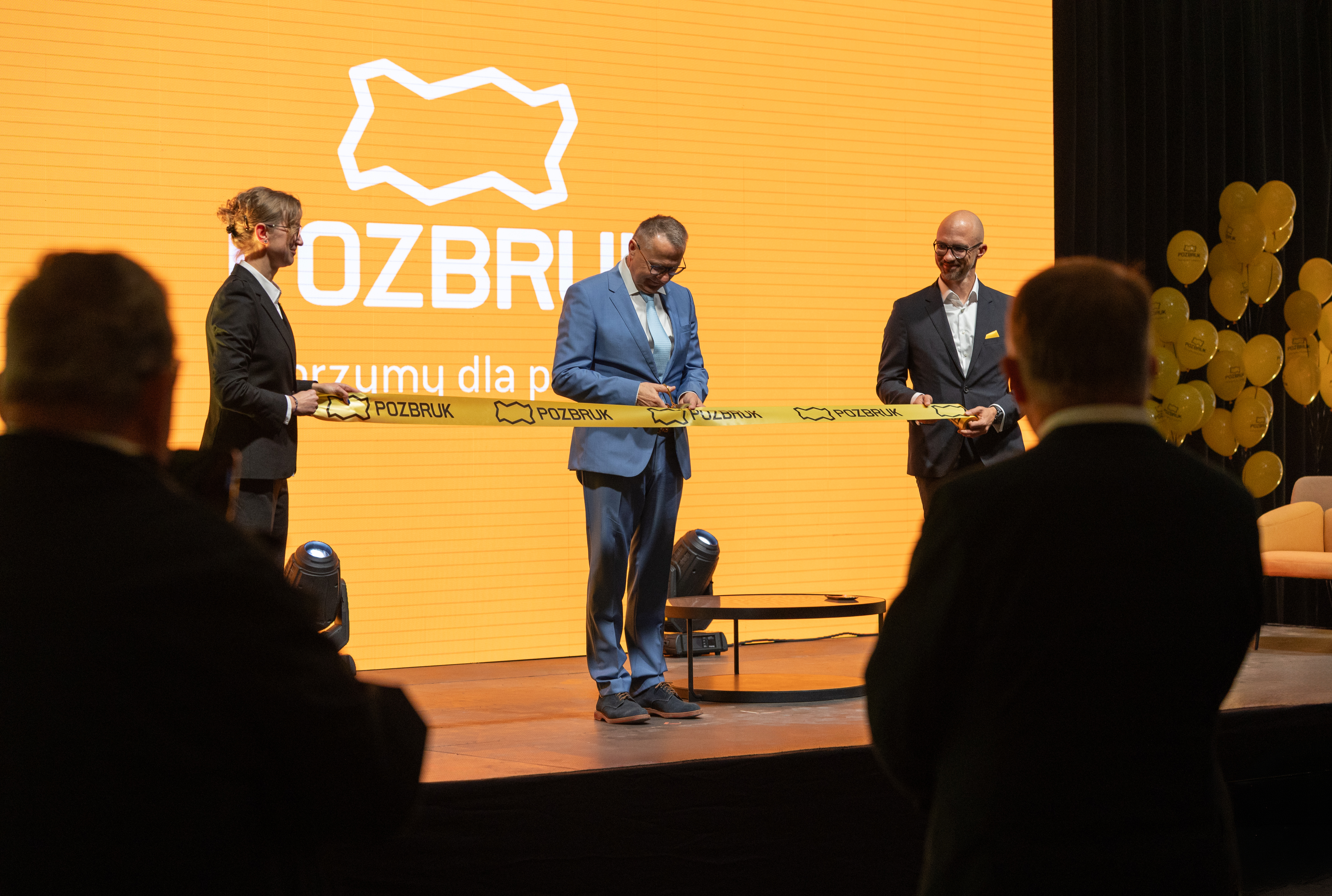Mężczyzna w niebieskim garniturze przecina żółtą wstęgę na scenie przed pomarańczowym ekranem z logo POZBRUK.