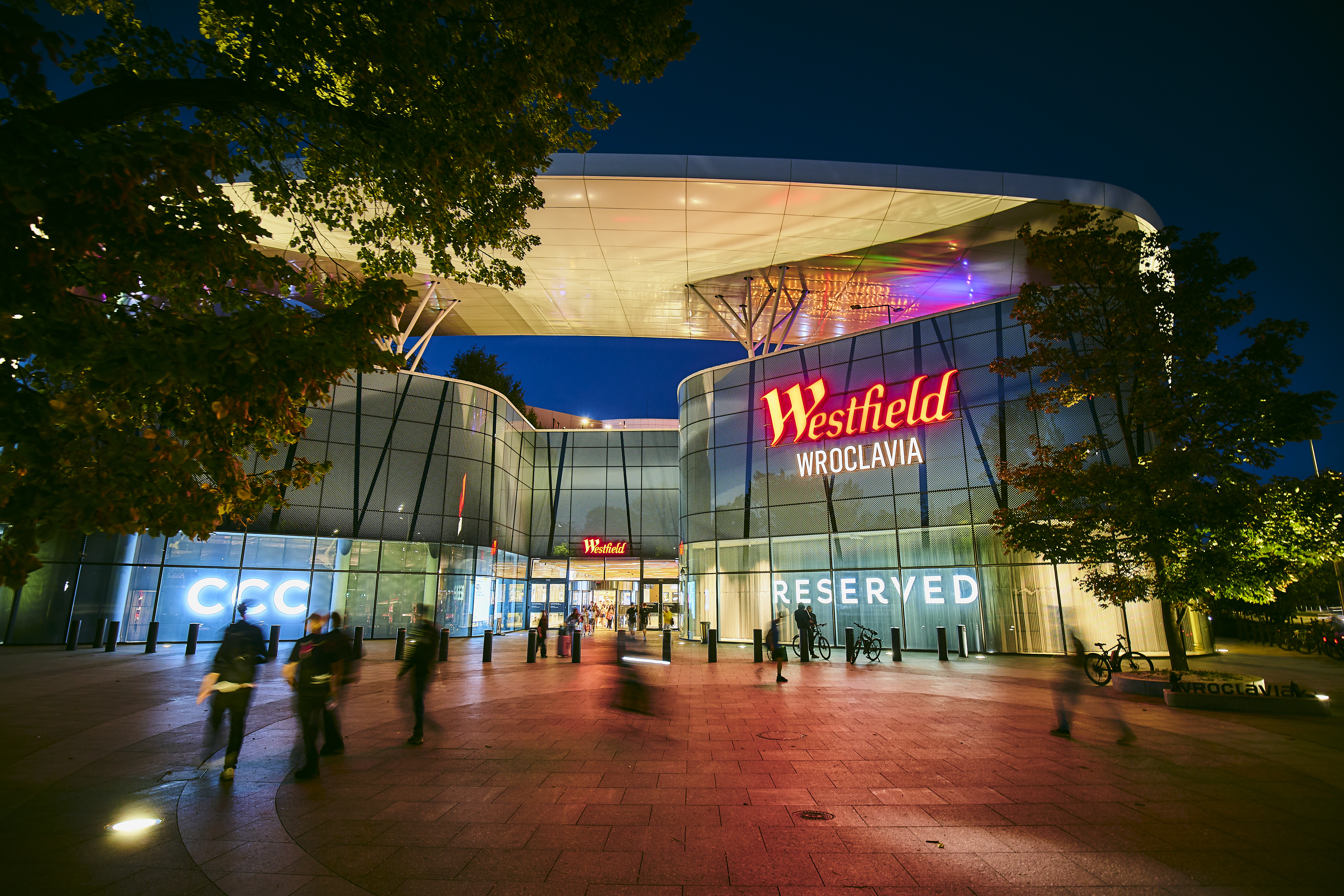 Nowoczesna fasada galerii Westfield Wroclavia z przeszkloną elewacją i białym, obłym zadaszeniem widoczna po zmroku, z rozmyciem przechodniów.