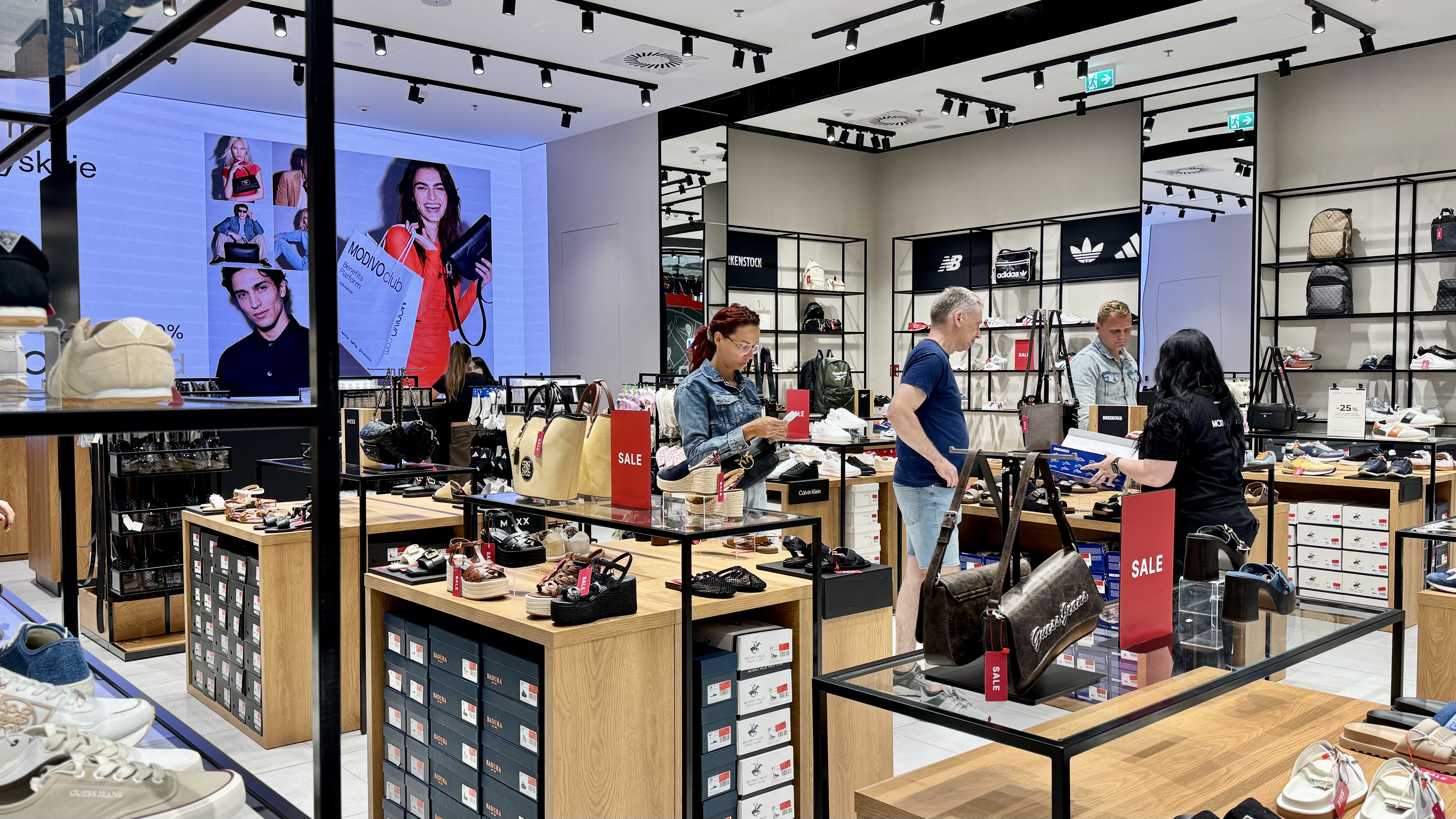 Nowoczesny salon obuwniczy z regałami pełnymi butów, torebek i plecaków, z klientami oraz pracownikami, drewnianymi ladami z produktami SALE i ekranem reklamowym.